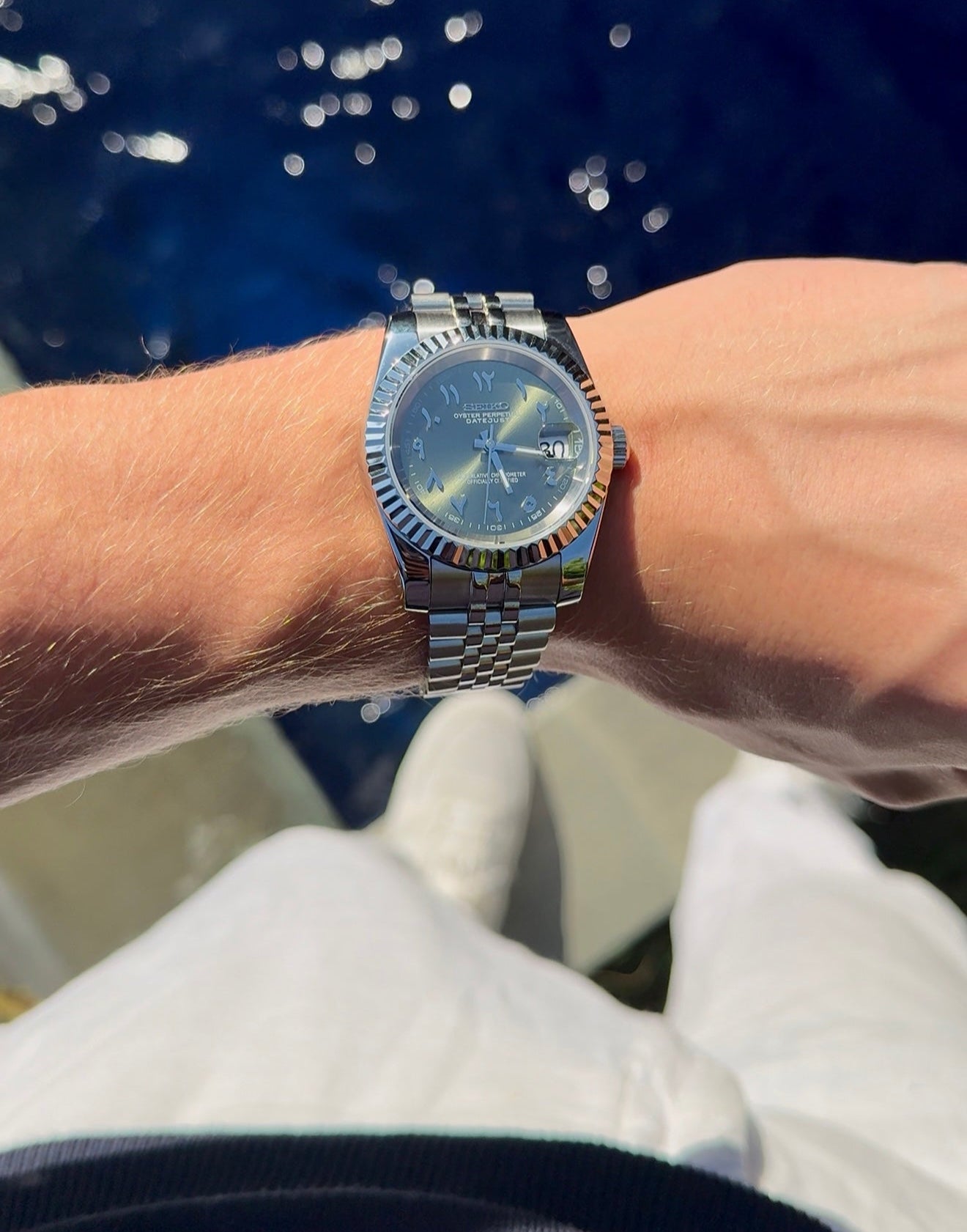 Close-up of a wrist wearing a silver watch with a blue face against a blurred water background