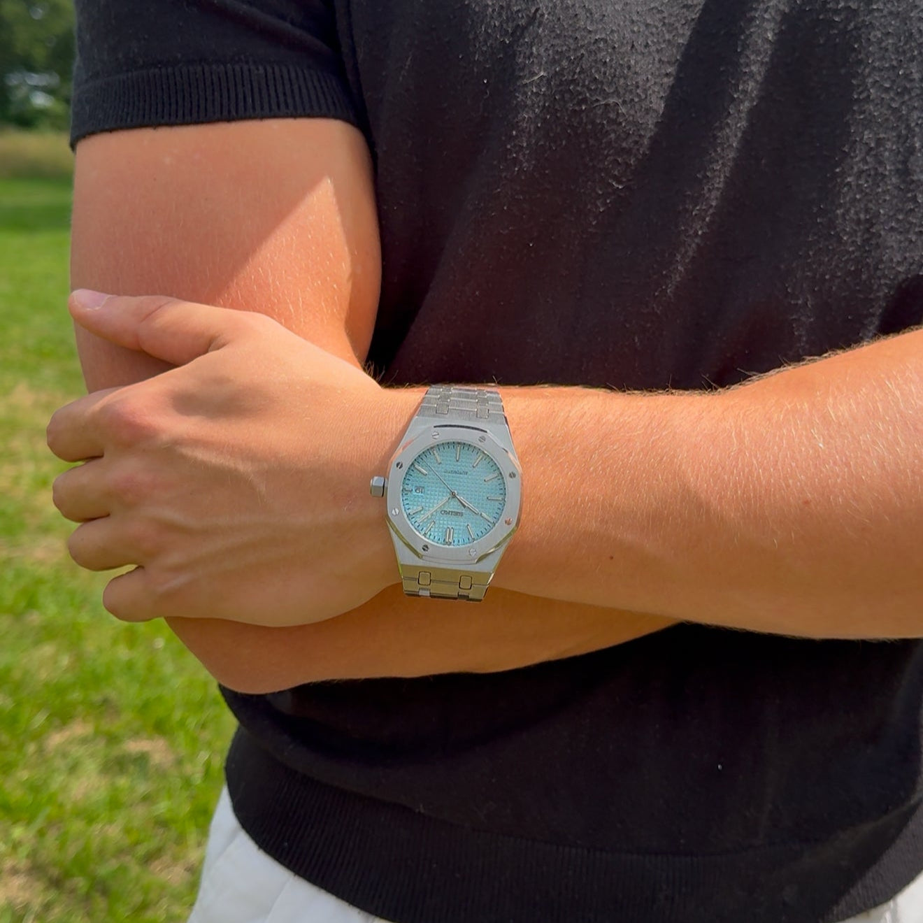 Person wearing a black polo shirt and light-colored pants with a silver watch on a grassy background